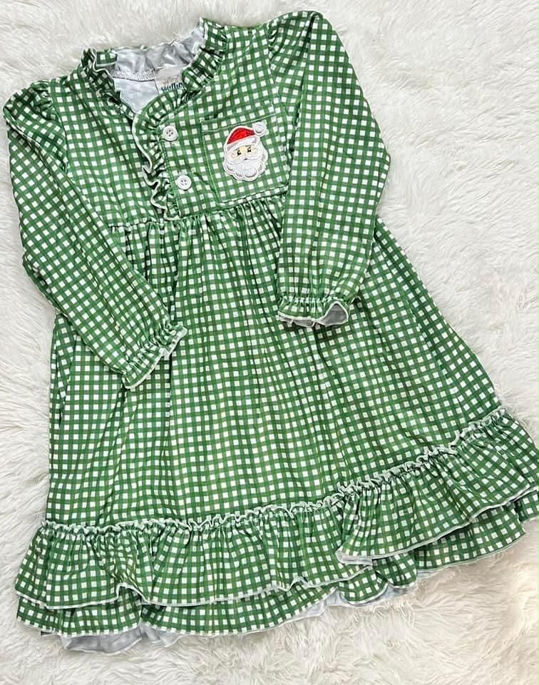 A green and white gingham long-sleeve baby dress with a ruffled collar, buttoned neckline, and a Santa face patch on the chest, laid flat on a white faux fur background.