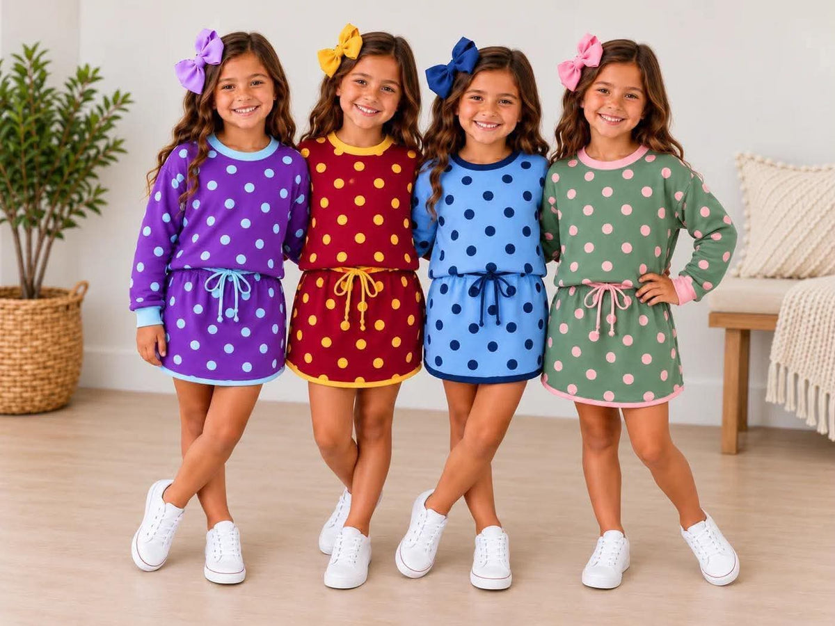 Four young girls standing together in a bright indoor room, each wearing a long-sleeve polka-dot dress in a different color: purple, red, blue, and green. The dresses have large polka dots, drawstring waist ties, and white sneakers.