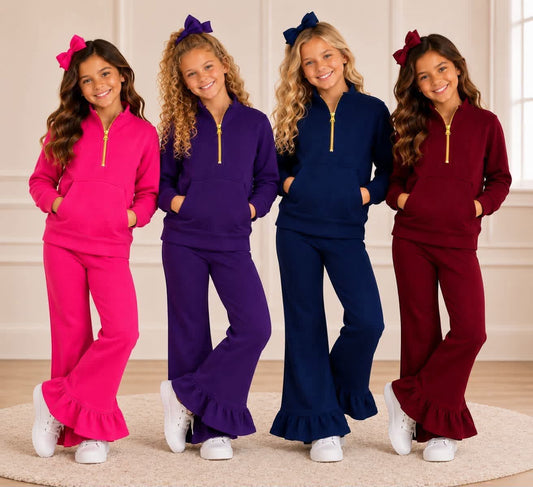 Four young girls modeling matching fleece tracksuits in bright pink, purple, navy blue, and burgundy, each with a half-zip top and flared pants, standing together in a bright indoor room.